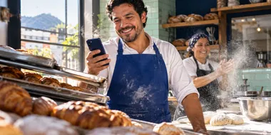 Panadero revisando su celular mientras su compañera hornea pan en su panadería
