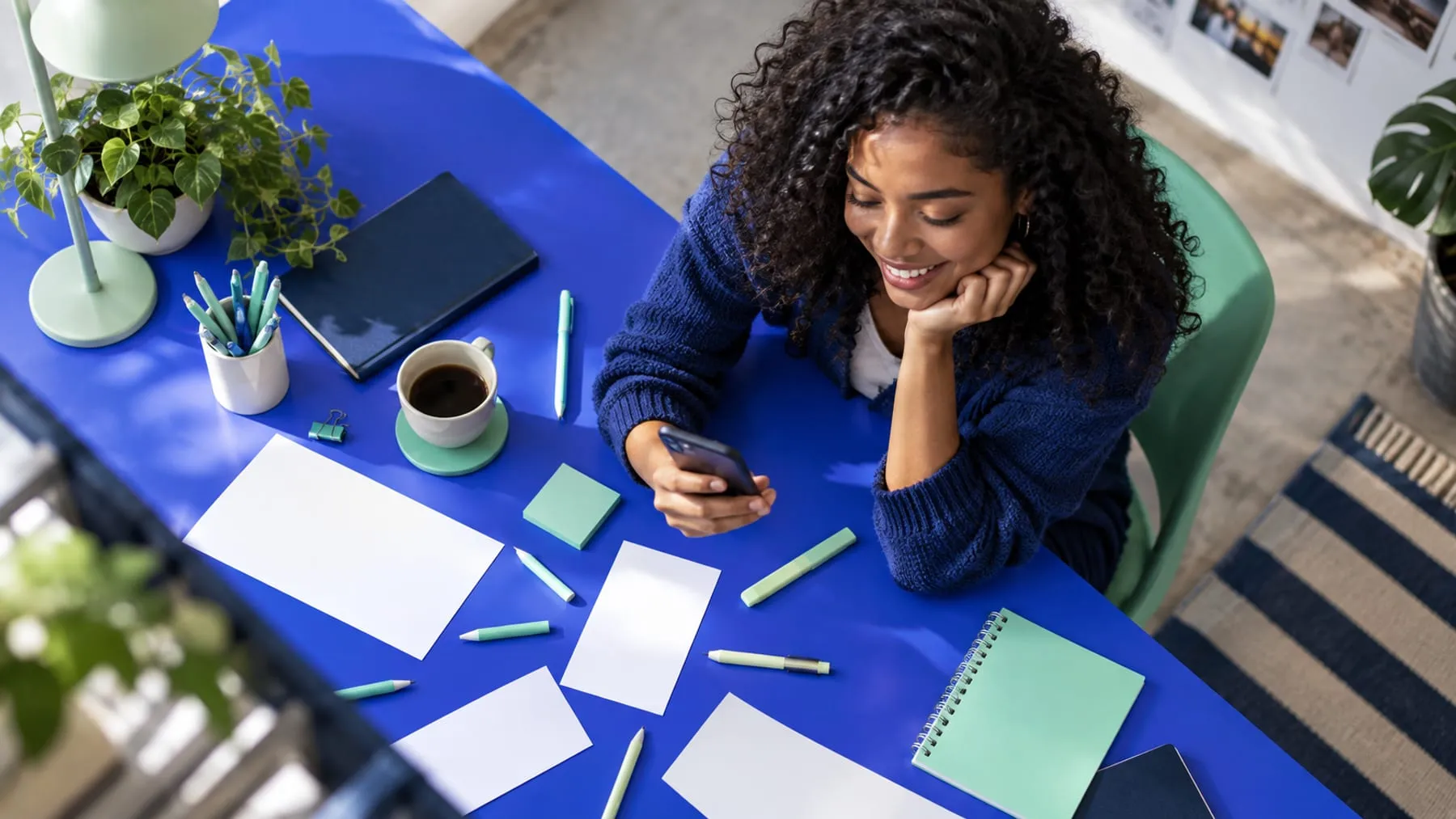 Vista superior de una mujer sonriendo mientras revisa su celular en un escritorio con plantas, café y artículos de oficina en tonos azul y menta
