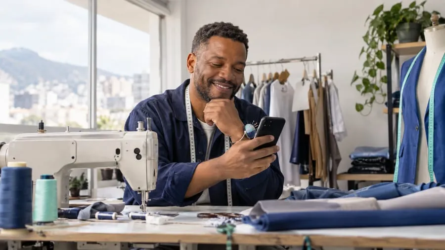 Sastre sonriendo mientras revisa su celular en su taller, con una cinta métrica al cuello